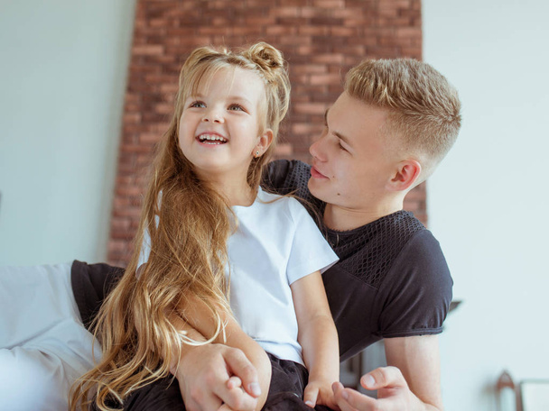caucasian siblings - teenager boy brother and little girl sister sitting on a couch (sofa) in modern loft interior - Photo, Image