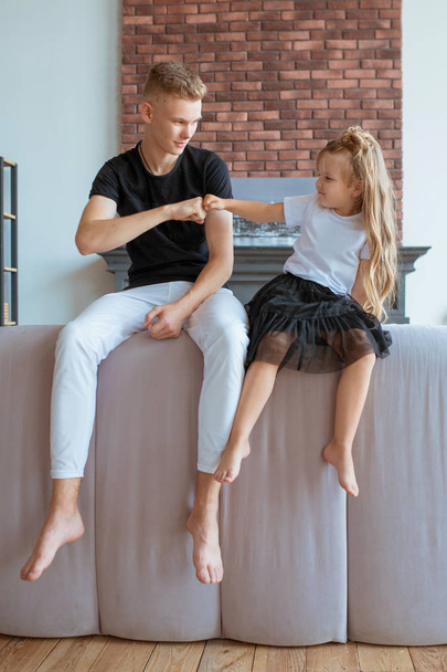 caucasian siblings - teenager boy brother and little girl sister sitting on a couch (sofa) in modern loft interior - Foto, imagen