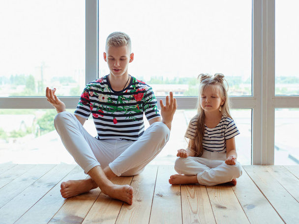hermanos caucásicos (hermano y hermana) haciendo yoga en casa junto a la ventana. Familia, parientes, relación y concepto deportivo. - Foto, imagen