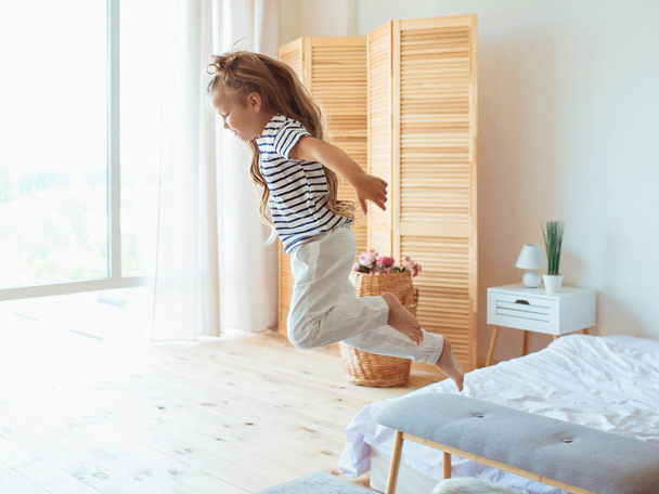 Маленькая кавказская девочка выпрыгивает из кровати в спальне. Счастье, сила, мотивация и утренняя концепция
 - Фото, изображение