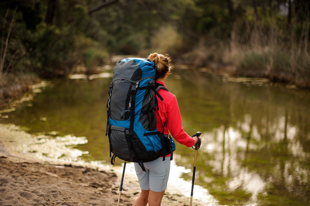 Rear view girl standing with hiking backpack and looking at the pond - Фото, зображення
