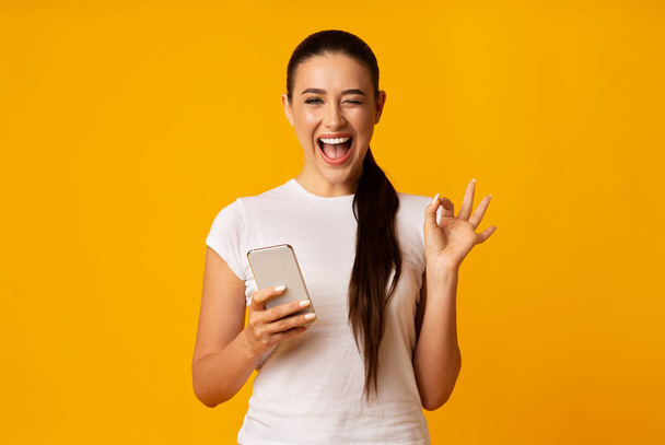 Великолепная женщина Жест ОК и держа сотовый телефон на желтом фоне
 - Фото, изображение