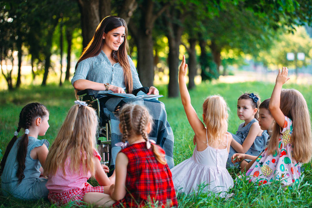 Vammainen opettaja tekee oppitunnin lasten kanssa luonnossa. Pyörätuolissa istuvan opettajan vuorovaikutus opiskelijoiden kanssa. - Valokuva, kuva
