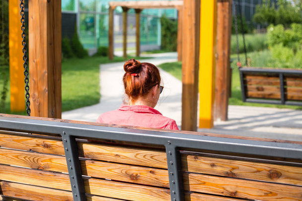 Όμορφη νεαρή γυναίκα με κόκκινα μαλλιά που κάθονται σε ένα παγκάκι σε ένα πάρκο της πόλης, χαλαρό κορίτσι - Φωτογραφία, εικόνα