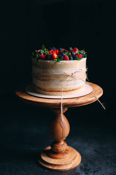 Fruit cake. Cake decorated with berries on a wooden stand on a black background. - Foto, afbeelding