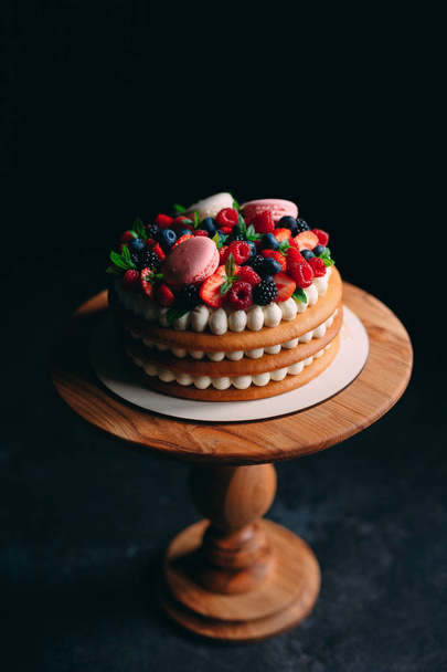 Fruit cake. Cake decorated with berries on a wooden stand on a black background. - Фото, изображение