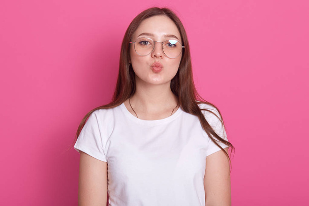 Impresionante modelo femenino posando con expresión de cara de beso en redondo rosado. Primer plano retrato de la elegante chica europea de pie frente a la pared del estudio, vestido casual camiseta blanca y gafas
. - Foto, Imagen
