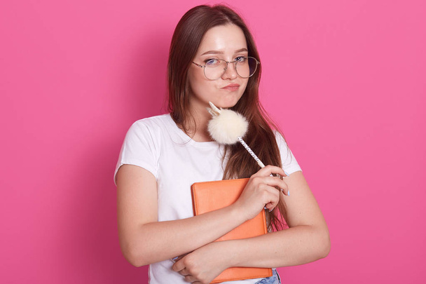 Horizontal shot of young woman with staright hair holding notebook and pen while standing against pink studio wall, looking directly at camera with thoughtful facial expression. Students concept. - Foto, Bild
