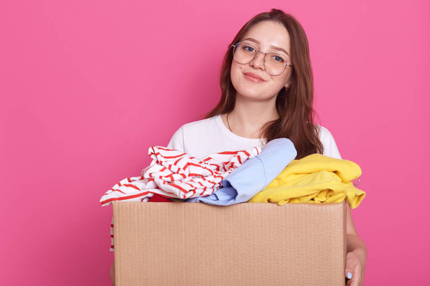 Horizontal shot of smiling female posing isolated over pink background and holding box with rausable clothes, clothing for children`s home or poor people, charming girl woman making charity. - Fotografie, Obrázek