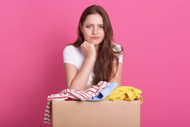 Close up portrait of beautiful woman with pensive facial expression, posing near box with old clothes, decides whom to give things for secondary use, standing over pink background. Donation concept. - Foto, imagen