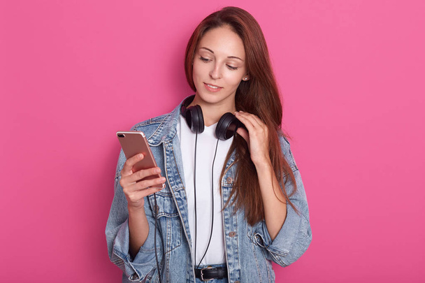 Young attractive girl standing isolated over pink background, holding her smartphone in hand, having headphones around neck and touches it with her fingers, looks at her device, wearing stylish outfit - Fotoğraf, Görsel