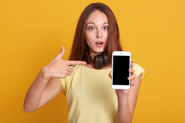 Studio sho of adorable woman showing phone with blank screen and pointing on it with her index finger, has astonished facial expression, keeps mouth opened, wearing yellow casual t shirt. Copy space. - Foto, imagen
