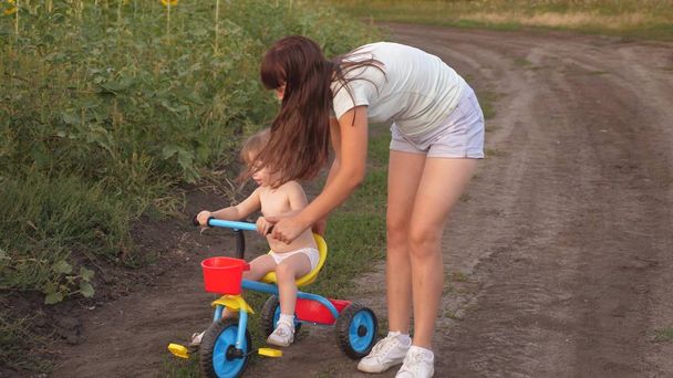 Mamá enseña a su hija a andar en bicicleta. La hermana juega con un niño pequeño. El concepto de la infancia feliz. un niño pequeño aprende a andar en bicicleta
. - Foto, imagen
