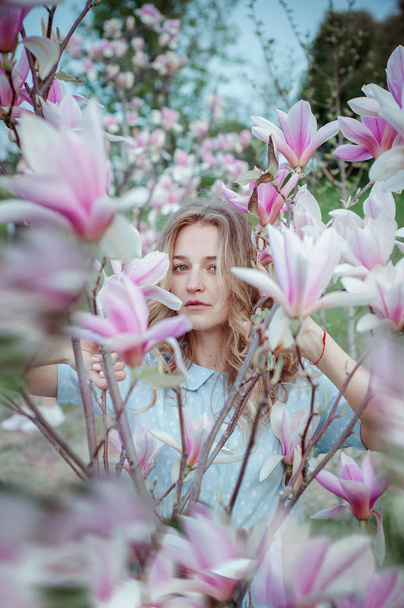 Beautiful happy young woman enjoying smell in a flowering spring garden.Beutiful tree magnolias,big flowers. Blonde with blue eyes and blue dress. - Φωτογραφία, εικόνα