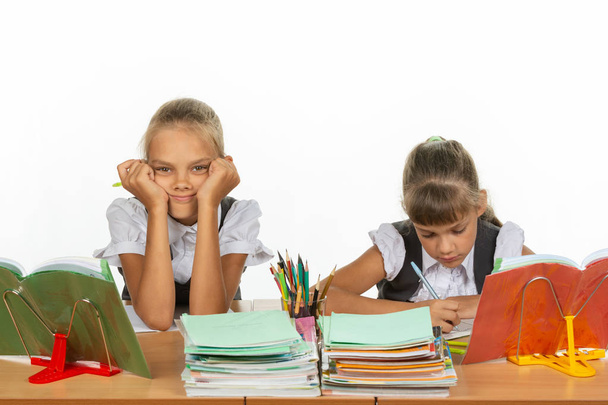 zwei Schulmädchen an einem Schreibtisch, die eine stützt sich auf ihre Hände und schaut in den Rahmen, die andere schreibt - Foto, Bild