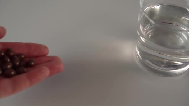 Dłoń rozlewa garść ciemnych brązowych kapsułek medycznych na szarym stole obok szklanki wody. Przygotowanie do podjęcia witamin i suplementów diety - Materiał filmowy, wideo