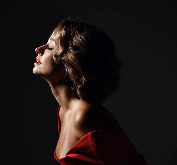 Beautiful trendy woman with curly hair and emotions in red dress posing on dark  - 写真・画像