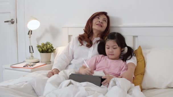 Tablet kullanarak Asyalı büyükanne evde torunu için masal okuyun. Üst düzey Çinli, büyükanne mutlu gece konsepti evde yatak odasında yatakta yatmadan önce genç kız dinlenmek. Yavaş çekim. - Video, Çekim