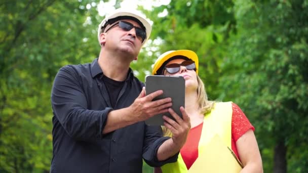 Dva absorbované stavební inženýry, muž a žena, které nosí ochranné přilby, které kontrolují staveniště pomocí digitálního tabletového počítače - Záběry, video