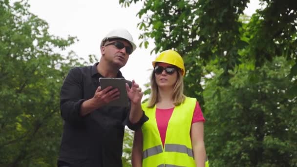 Dwóch wchłoniętych inżynierów budowlanych, mężczyzny i kobiety, noszących Hełmy ochronne kontrolują miejsce budowy za pomocą cyfrowego komputera typu Tablet - Materiał filmowy, wideo