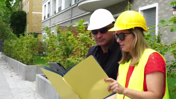 2人の吸収された建設技術者、男性と女性、デジタルタブレットコンピュータを使用して建設現場を検査する保護ヘルメットを着用 - 映像、動画