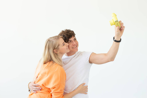 面白いクレイジー若いカップルのブロンドの女の子と白い背景にポーズヴィンテージ黄色のフィルムカメラに自分撮りを取るヒップスターの男。写真趣味の概念. - 写真・画像