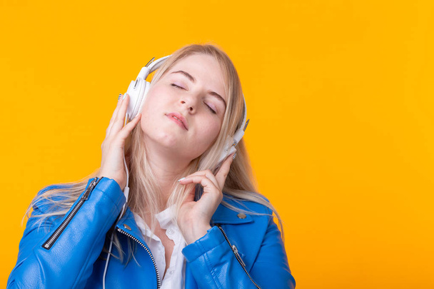 Portrait of pretty young girl blonde female student holding smartphone with blue leather jacket headphones posing on a yellow background. Concept of listening to online radio and music subscription. - Fotografie, Obrázek