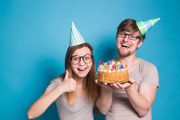Αστείο φυτό άνθρωπος και γυναίκα φορούν αργίες καπέλα και γυαλιά κρατώντας τούρτα γενεθλίων με κεριά πάνω από μπλε φόντο - Φωτογραφία, εικόνα