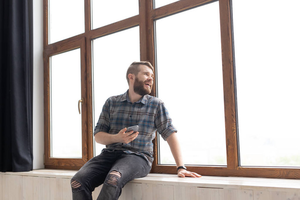 Jonge knappe man hipster zittend op de vensterbank in de buurt van een groot raam met een smartphone in zijn handen en kijken naar de bouwplaats thuis. Beginner architect concept. - Foto, afbeelding