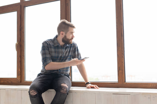 Young handsome man hipster sitting on the windowsill near a large window with a smartphone in his hands and looking at the construction site at home. Novice architect concept. - 写真・画像