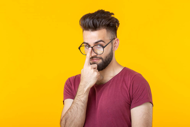 Jeune homme hipster pensif avec une barbe tenant ses lunettes debout sur un fond jaune. Le concept d'attention et de choix difficile
 - Photo, image