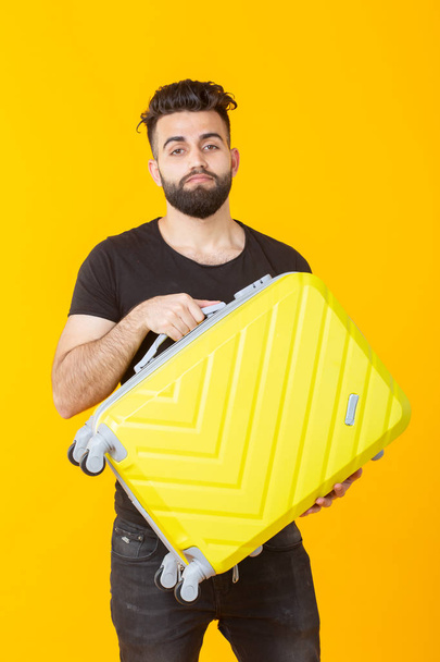 Sarı bir arka plan üzerinde elinde sarı bir bavul tutan sakallı sevimli güzel genç arap adam. Seyahat ve tatil kavramı - Fotoğraf, Görsel