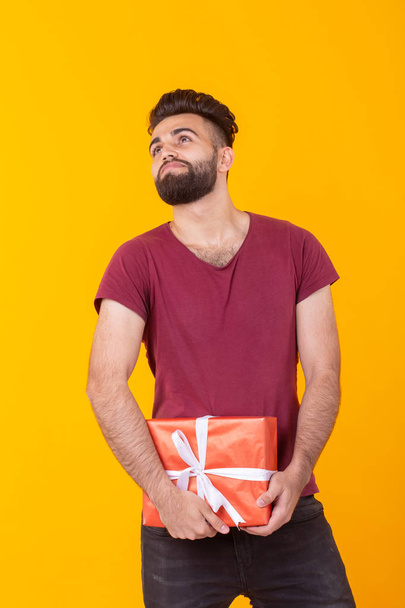 Cute handsome young hipster man in burgundy shirt holding a red gift box posing on a yellow background. Concept of holidays and birthdays - Photo, Image