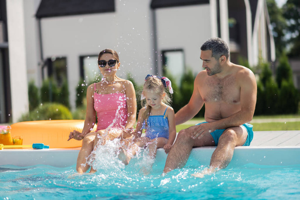 Daughter splashing water while sitting near pool with parents - Foto, imagen