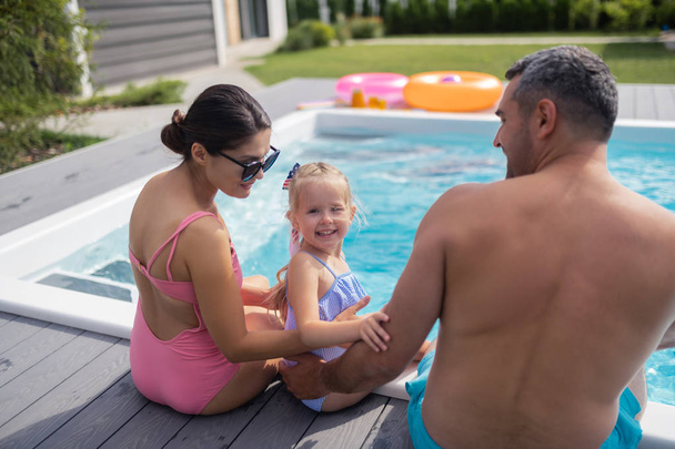 Daughter smiling while resting near pool with mom and dad - 写真・画像