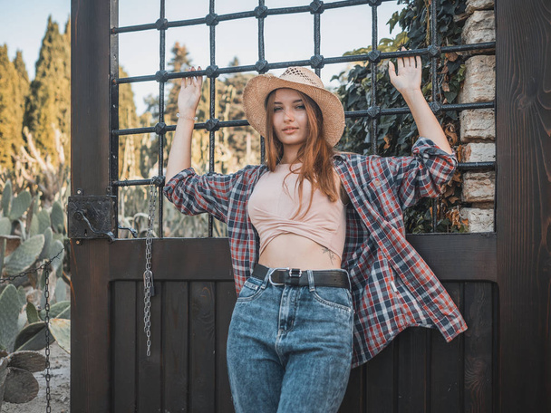 麦わら帽子をかぶった牧場の若い赤髪の女性、グリルの背景にチェック柄のシャツとジーンズ。カジュアルスタイル - 写真・画像