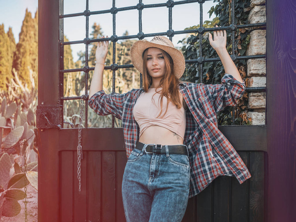 Young red-haired woman on a ranch in a straw hat, a plaid shirt and jeans on the background of the grill. Casual style - 写真・画像