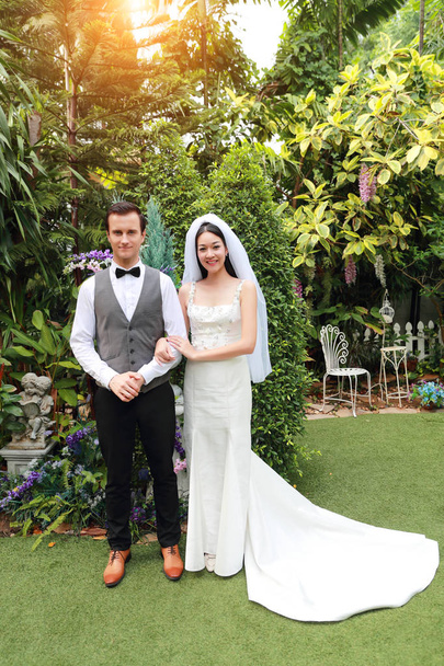 guapo caucásico novio está sosteniendo su asiática novia mano con amor y sonriendo cara durante boda o pre boda foto pose mientras de pie en jardín
 - Foto, imagen