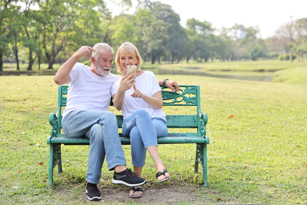 senior caucasian grandfather and grandmother sitting in park while using video call from smart phone to grandchildren with happy face and laughing  - Photo, Image