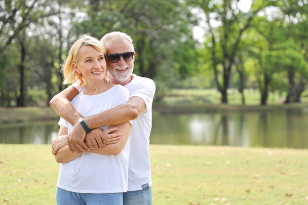 elderly caucasian couple grandfather and grandmother with white shirt, blue jean and sun glasses standing and embracing in park during summer time on wedding anniversary day - Photo, Image