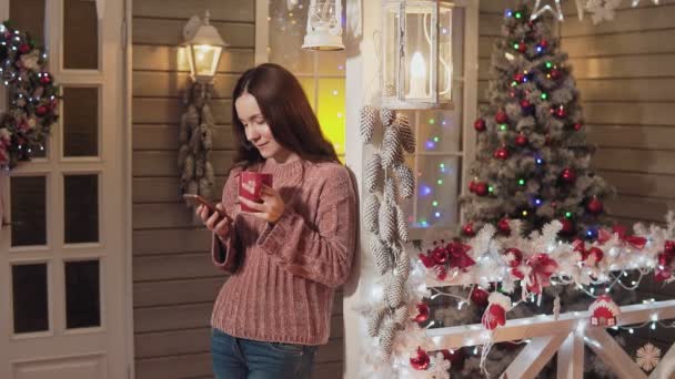 Young girl with red cup of tea or coffee and mobile phone. Xmas and new year concept. Brunette woman smiling drinks tea use phone with christmas tree and lights at background. Copyspace at right - Πλάνα, βίντεο