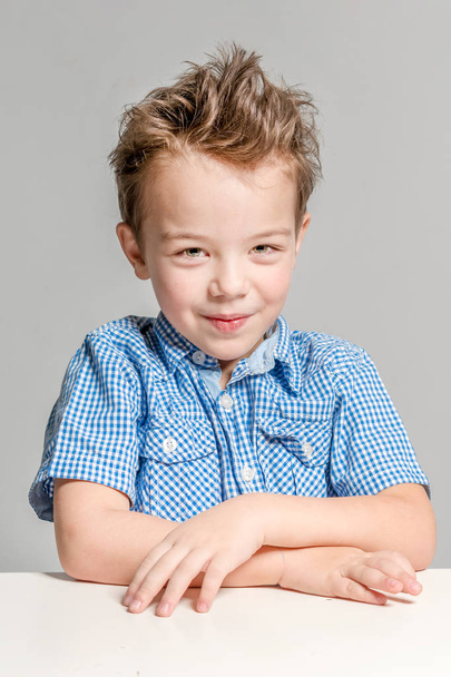 Cute boy in a blue shirt sitting at the table on a gray backgrou - Foto, afbeelding
