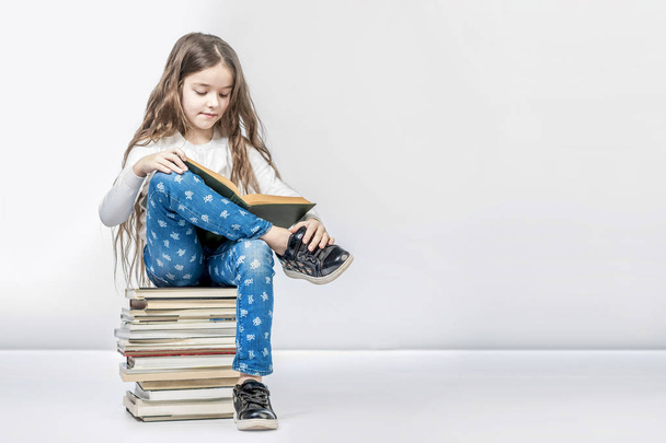Cute girl sitting on a stack of books and reading a book on a wh - Foto, afbeelding