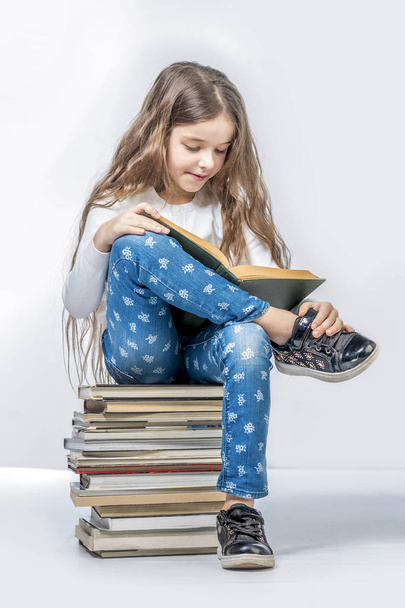 Cute girl sitting on a stack of books and reading a book on a wh - Fotografie, Obrázek
