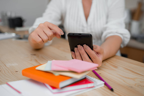 Young lady using a dark smartphone inside home. Woman holding a black cell phone in a house ambient. Girl with technological devices and office supplies with blank space. - Foto, Imagem