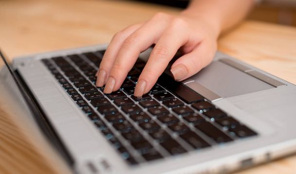 Młoda dama przy użyciu szarego komputera przenośnego i wpisując czarną klawiaturę z obiema rękami w pomieszczeniu. Materiały biurowe, urządzenia technologiczne i drewniane biurko. - Zdjęcie, obraz