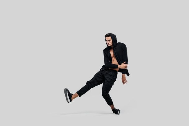Jeune beau jeune danseur vêtu de pantalon noir, un sweat-shirt sur un torse nu soulève une jambe tout en dansant la danse de rue
 - Photo, image