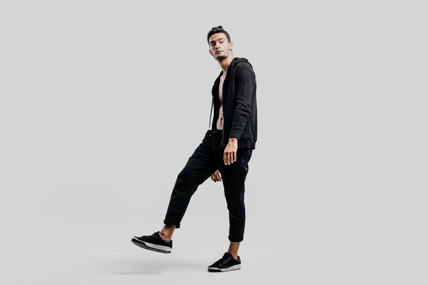 Young handsome dancer dressed in black pants and a sweatshirt on a naked torso stands on the white background - Photo, image