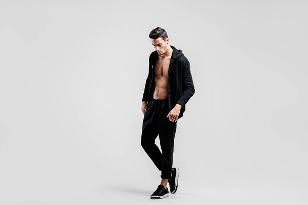 Young handsome dancer dressed in black pants and a sweatshirt on a naked torso stands on the white background - Foto, Imagen