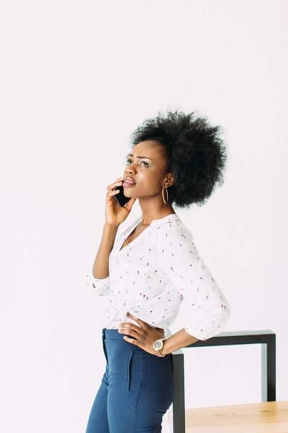 cheerful young afro american business woman talking on cell phone, standing near the modern chair, isolated on white - Fotografie, Obrázek